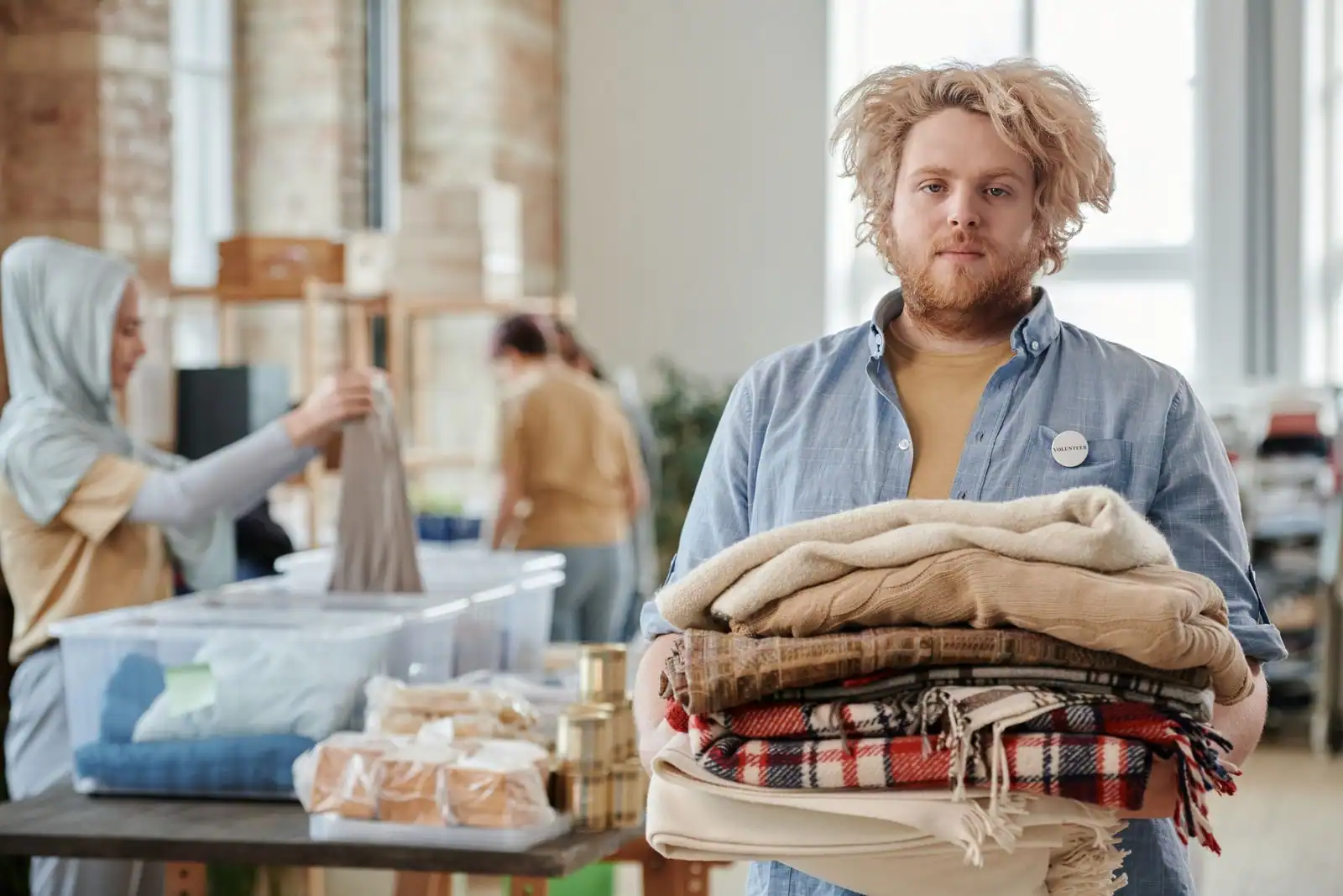 Homme tenant une pile de vêtements et plaids pliés lors d'un événement de vêtements de seconde main, personnes triant des habits en arrière-plan