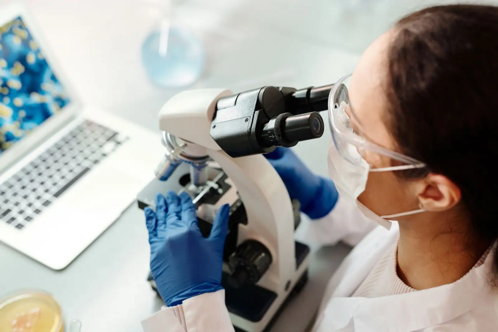 Une chercheuse en blouse et gants observe un échantillon au microscope dans un laboratoire de biologie.