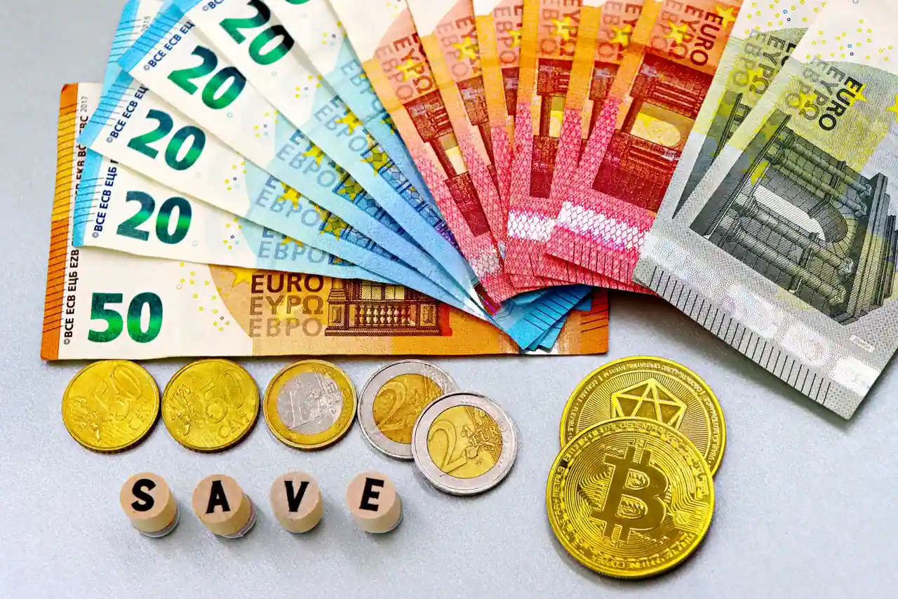 Billets et pièces en euros étalés sur une table claire, près d'une pièce dorée et de lettres en bois.