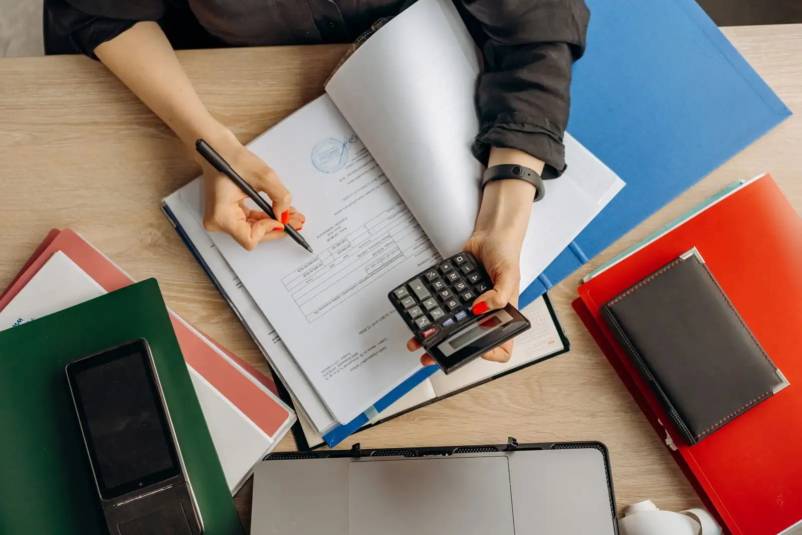 Personne examinant des documents comptables avec une calculatrice et un stylo, sur un bureau de travail.