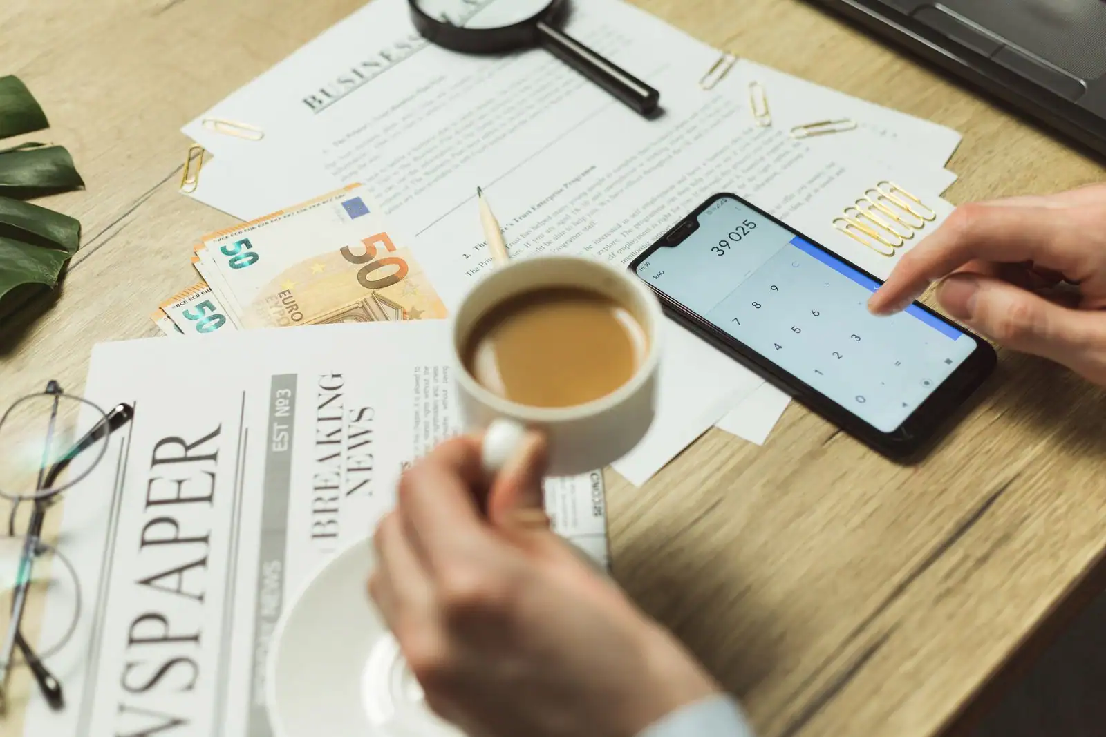 Des billets en euros, des documents et un smartphone affichant une application posés sur un bureau en bois, image illustrative.