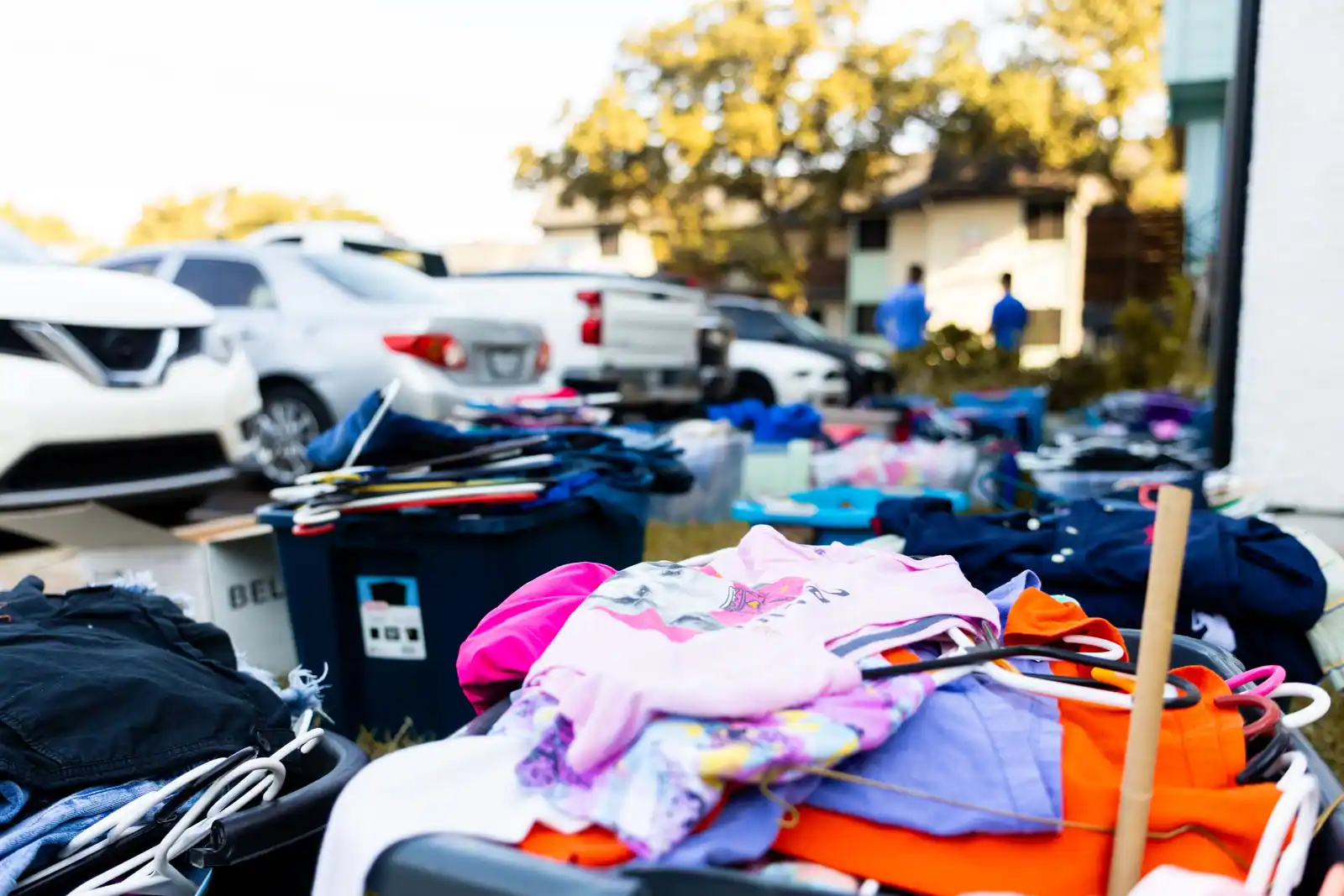 Vide-grenier en extérieur, vêtements et objets d'occasion étalés sur des bacs devant des voitures et une maison