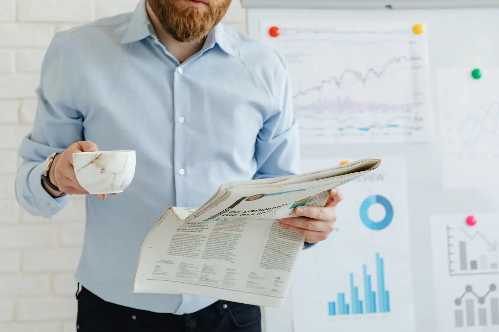 Un homme lit un journal économique, une tasse à la main, devant des graphiques financiers affichés au mur.