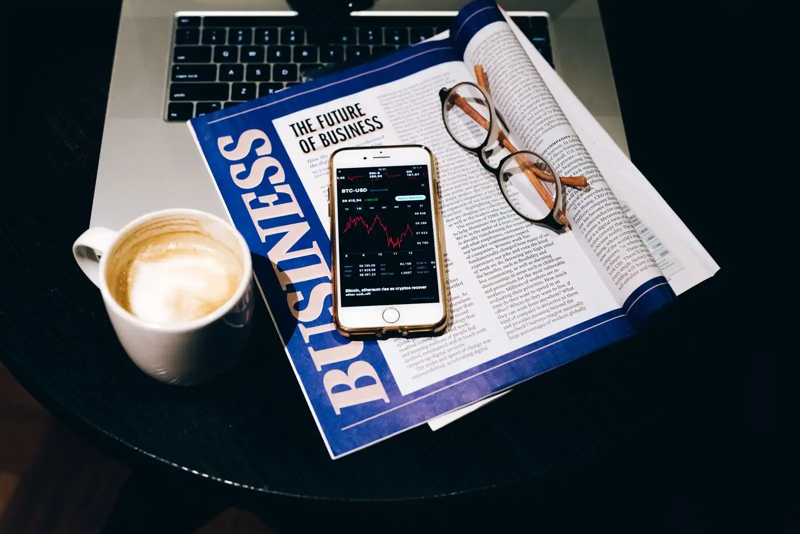 Magazine économique ouvert près d'un téléphone affichant un graphique de cours, des lunettes et un café