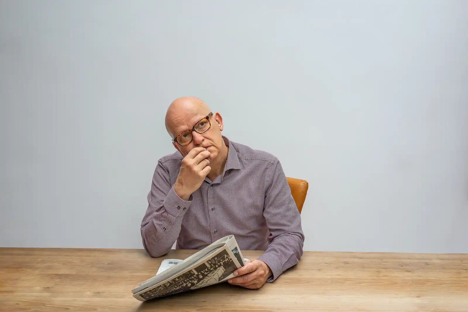 Un homme à lunettes suspend sa lecture, un journal plié devant lui sur une table en bois clair.