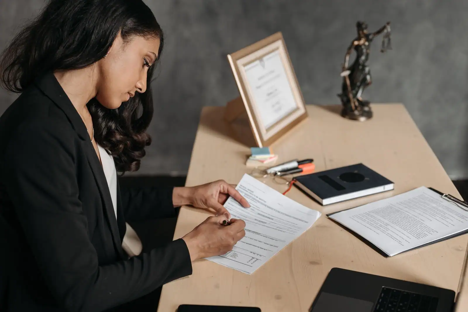 Une jeune femme en veste sombre annote un document juridique à son bureau, une statuette de la justice en arrière-plan