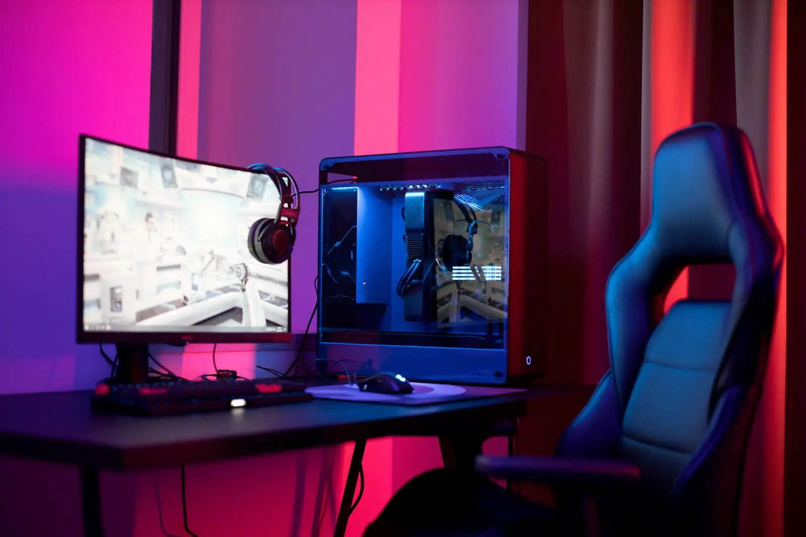 Écran affichant un jeu vidéo, tour d'ordinateur et fauteuil de gamer dans une pièce aux néons roses.
