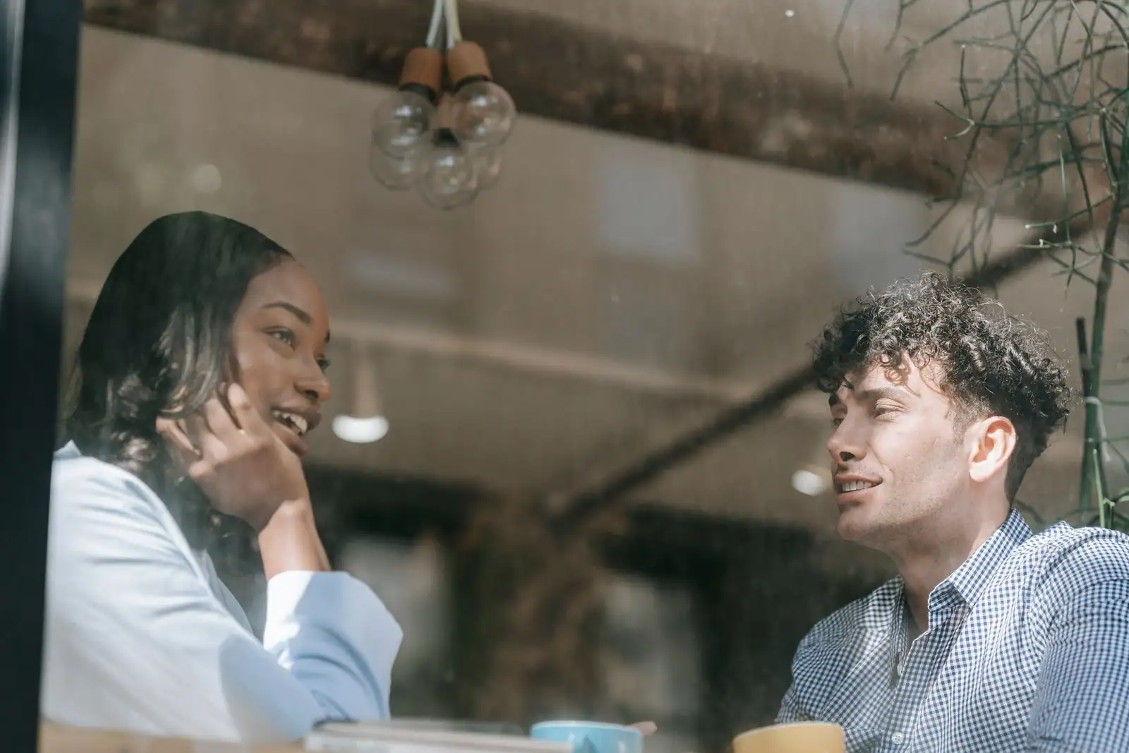 Deux personnes attablées se font face et discutent, vues à travers la vitre d'un café, tasses devant elles.