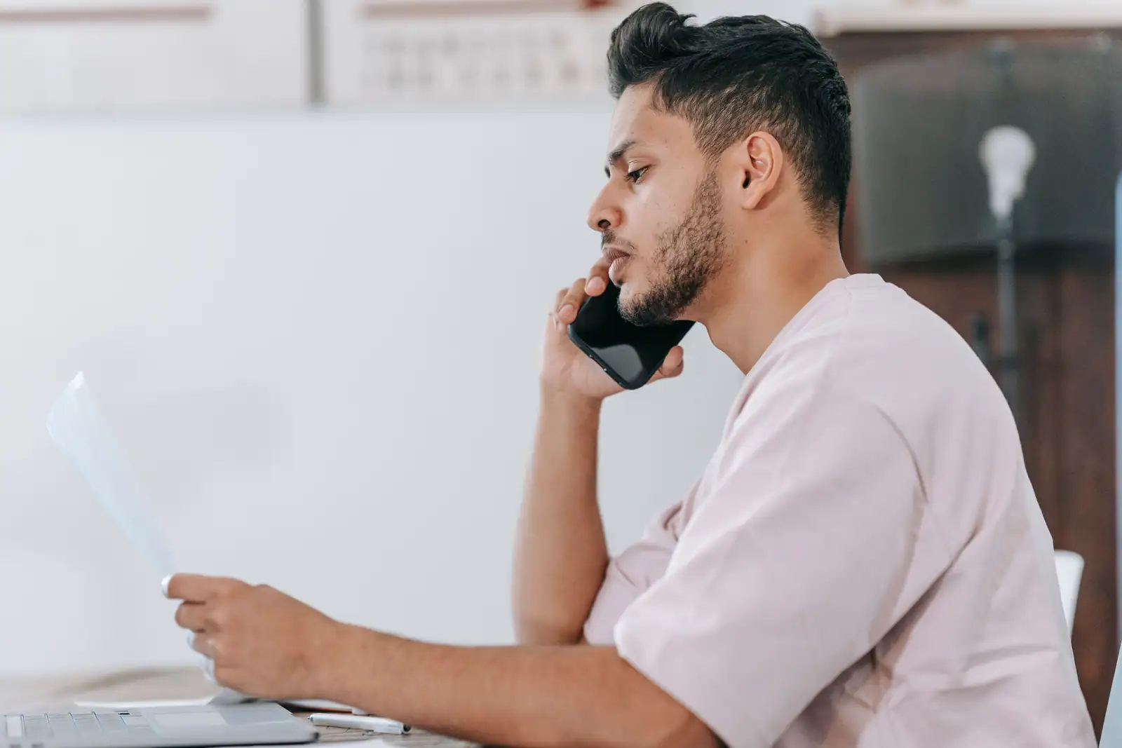 Homme au téléphone tenant un document imprimé devant son bureau, en train de joindre un service client
