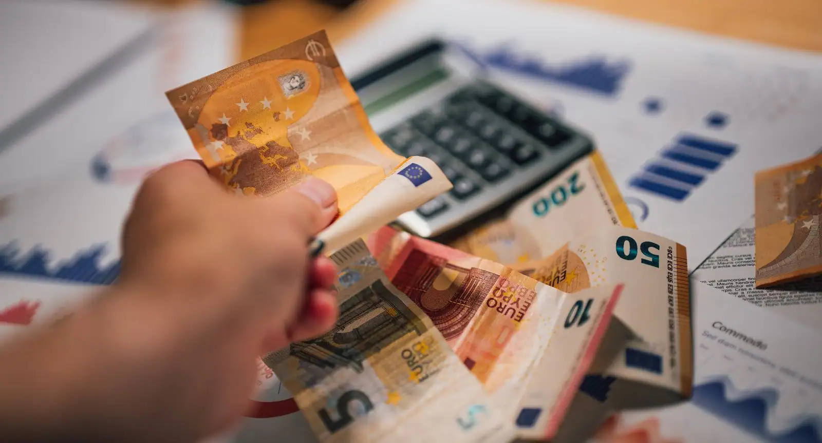 Main tenant plusieurs billets en euros au-dessus d'une calculatrice et de documents chiffrés.