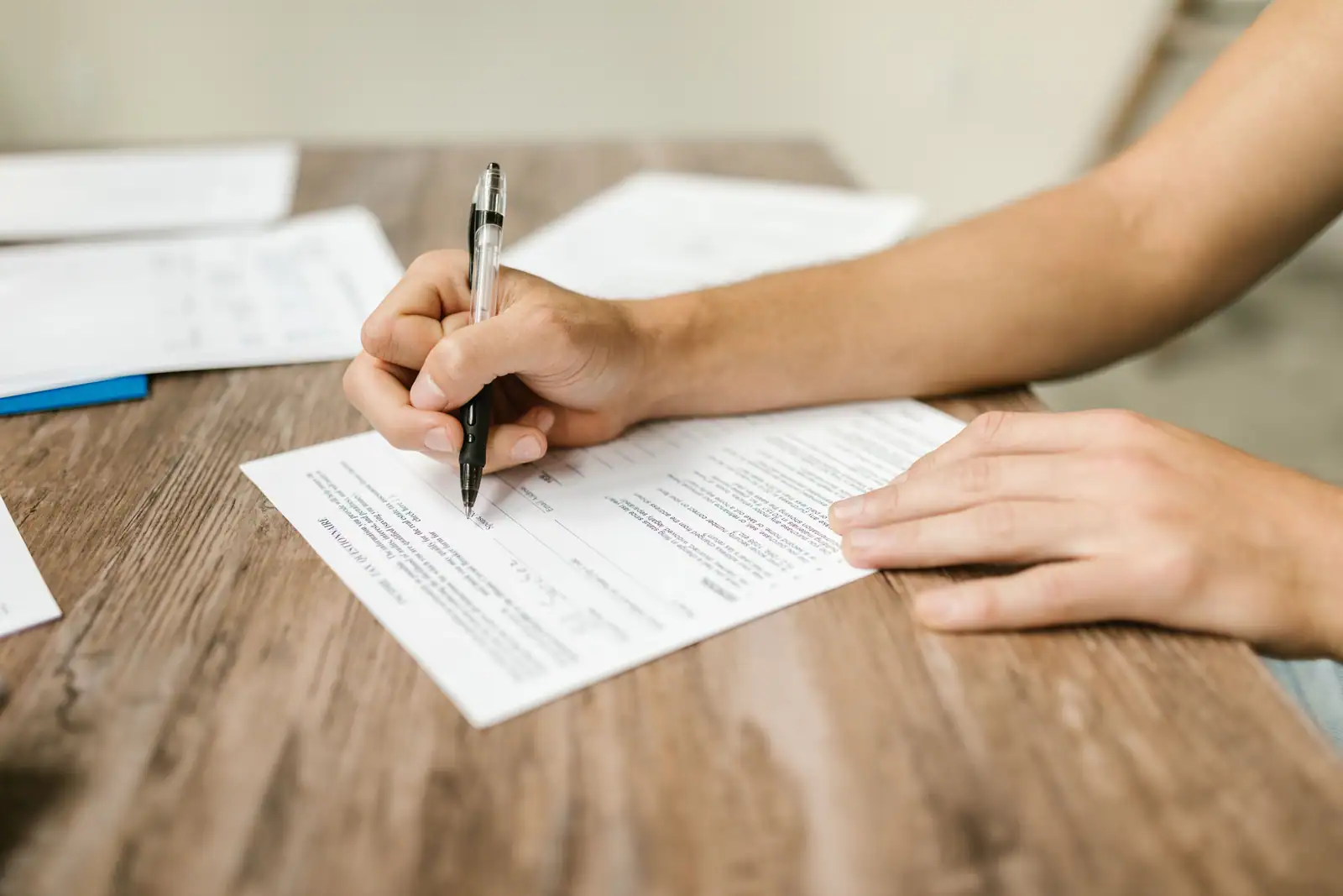 Mains remplissant au stylo un formulaire papier posé sur une table en bois, documents à côté