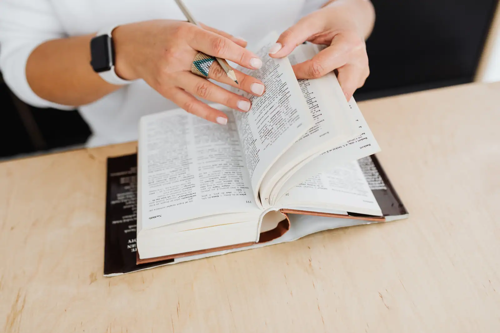 Mains feuilletant un gros dictionnaire ouvert sur une table claire, un crayon entre les doigts.