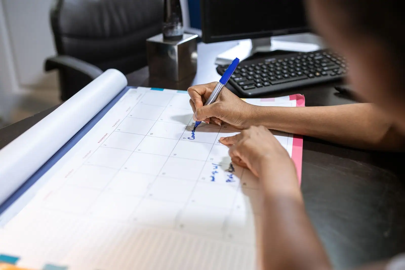 Une main annote au stylo bleu les cases d'un grand calendrier mensuel posé sur un bureau.