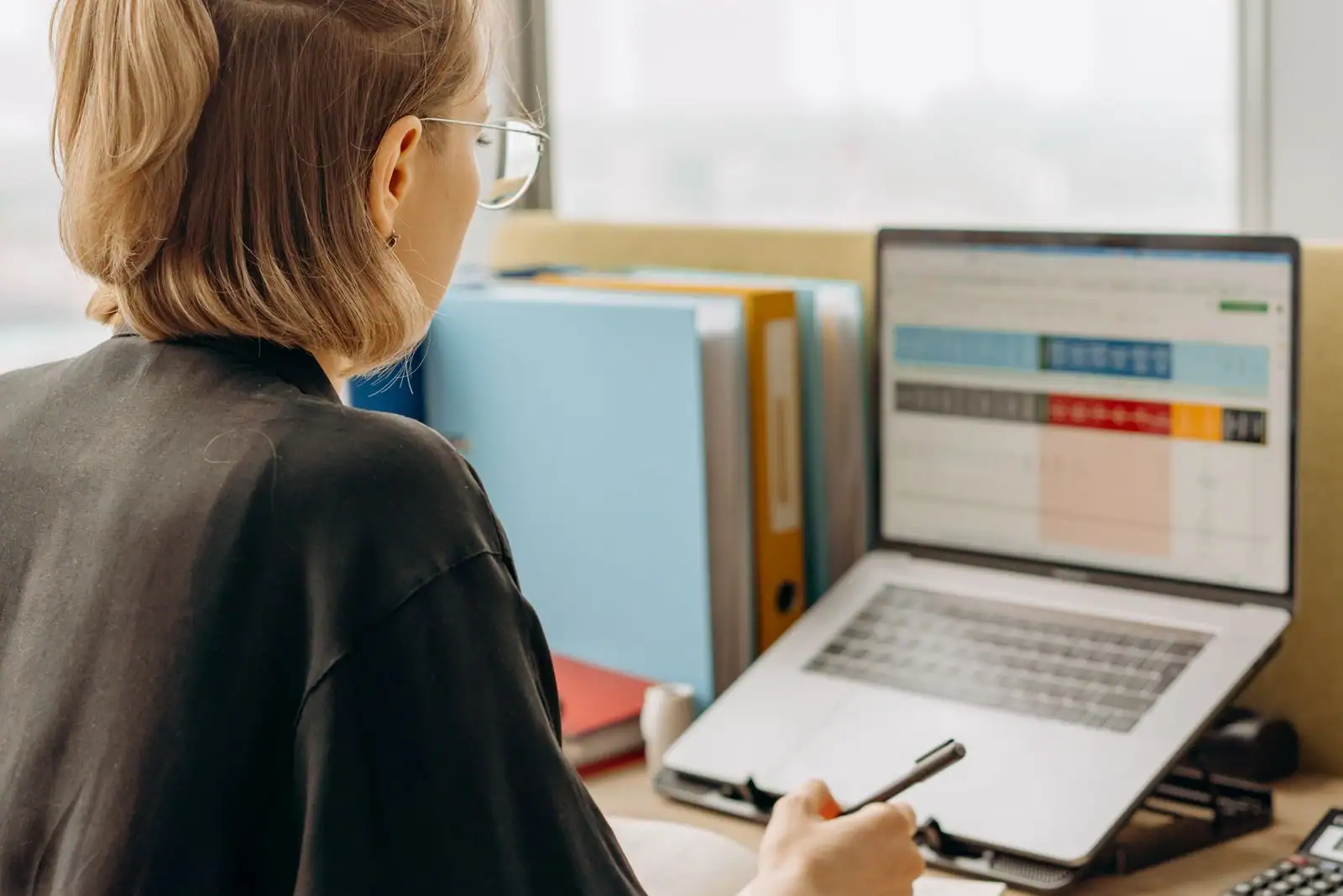 Personne de dos devant un ordinateur portable, classeurs à levier alignés sur un bureau administratif