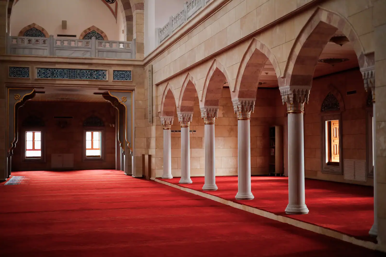 Colonnes blanches et arcades d'une salle de prière vide, tapis rouge au sol et lumière du jour.