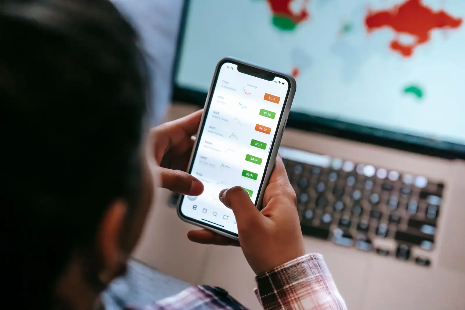 Personne consultant une application de suivi boursier sur smartphone, ordinateur portable en arrière-plan